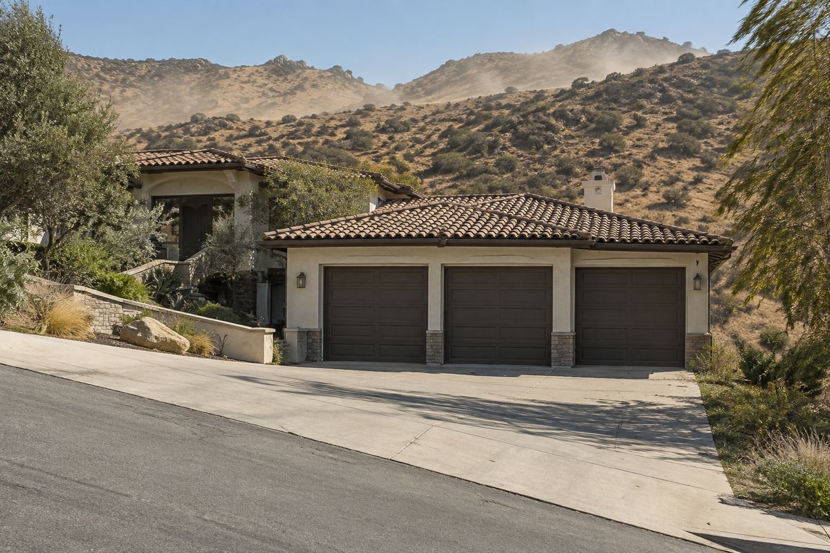 Hillside home with three garage doors and a sloped driveway below dry golden hills