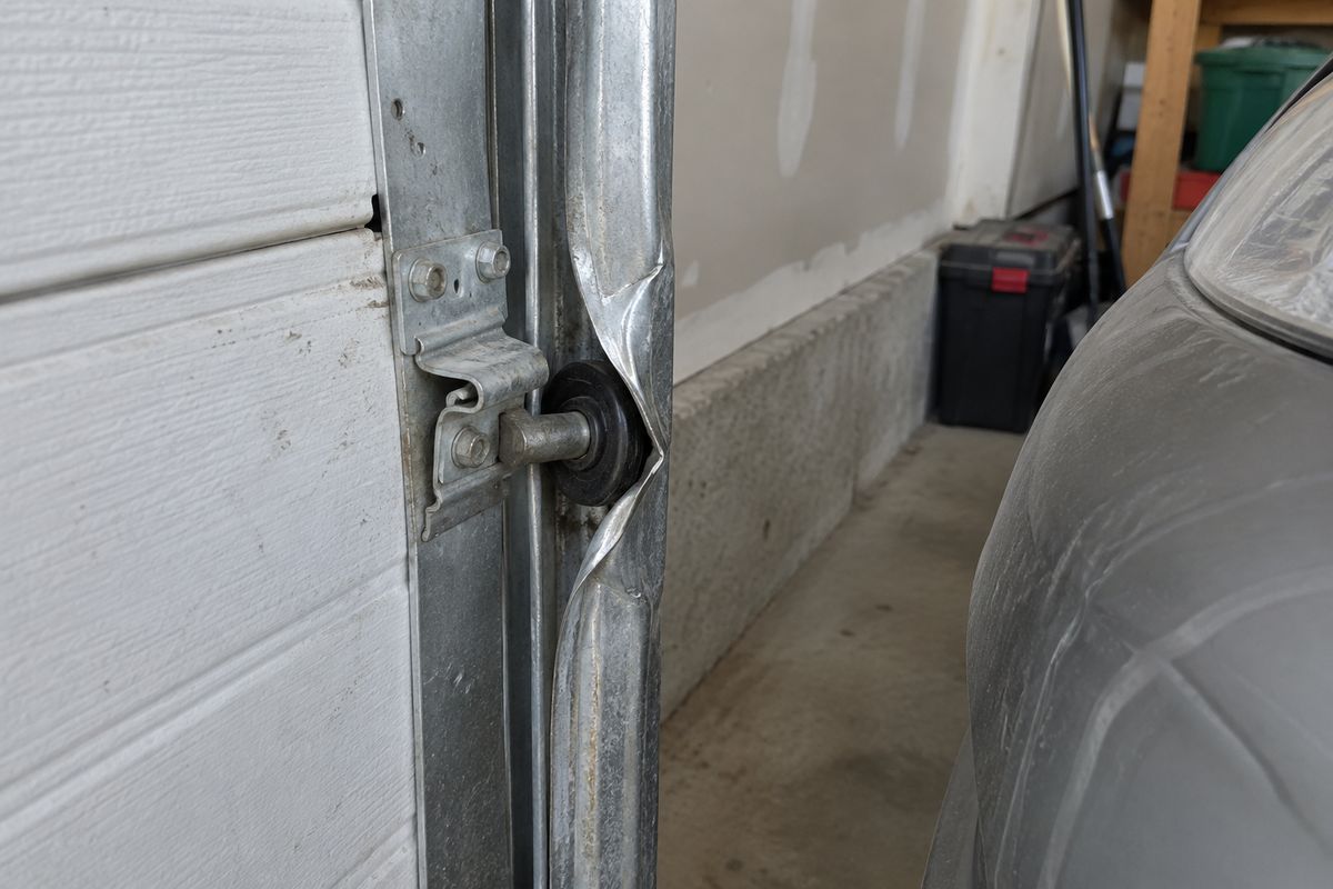Vertical garage door track creased and bent at bumper height, with a car parked behind