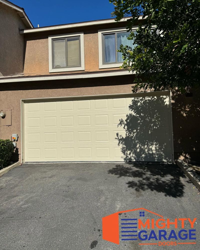 Cream raised-panel double garage door on a two-story stucco home with tree shadows across the driveway