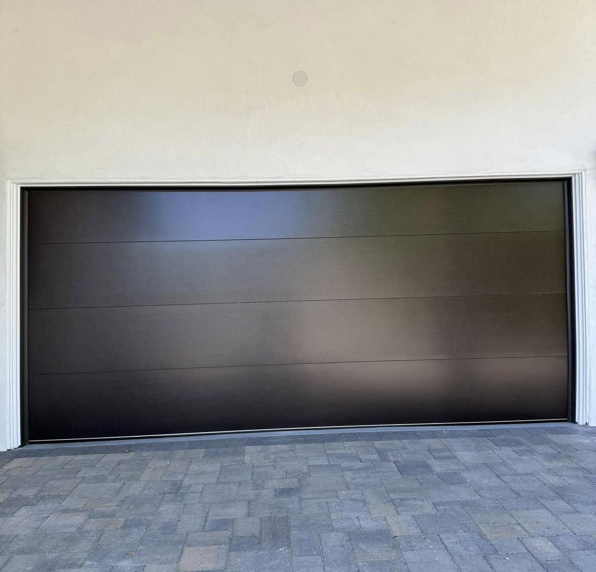 Wide dark bronze flush-panel garage door on a light stucco Los Angeles home with a gray paver driveway